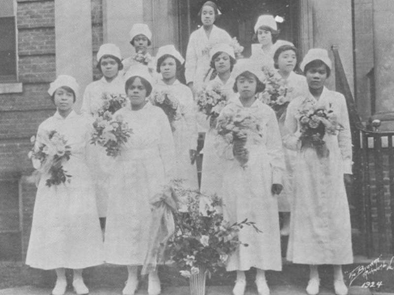 10 nurses dressed in commencement regalia and holding flowers in an archival photo from the nineteen twenties