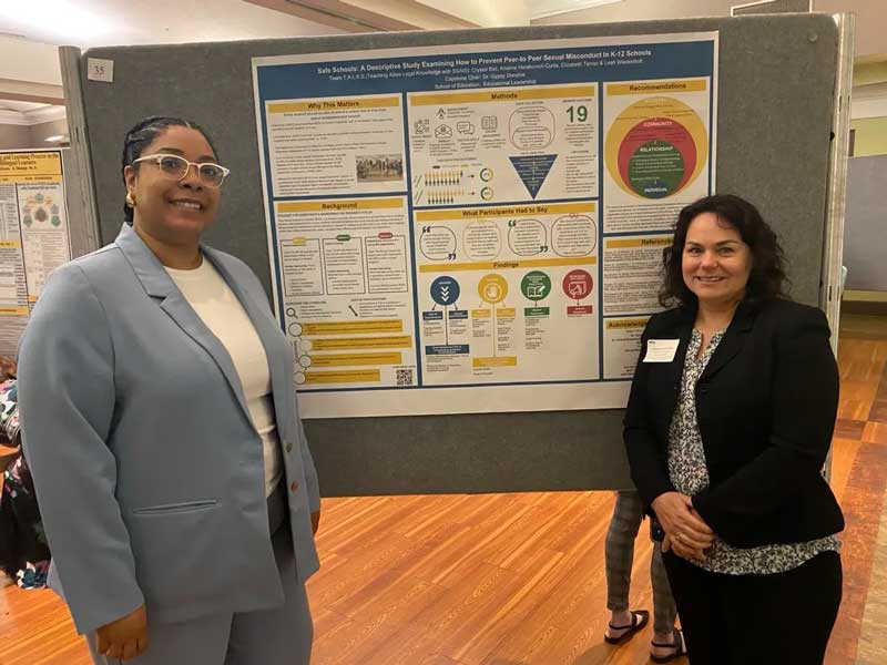 Crystal Bell and Kristine Herakovich-Curtis standing in front of a research poster titled 'safe schools: a descriptive study examining how to prevent peer-to-peer sexual misconduct in k-12 schools'