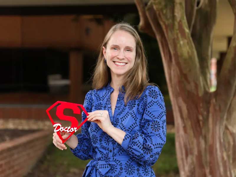 brittany craven smiling and holding a handcrafted wooden Superman shield with the word doctor across the middle