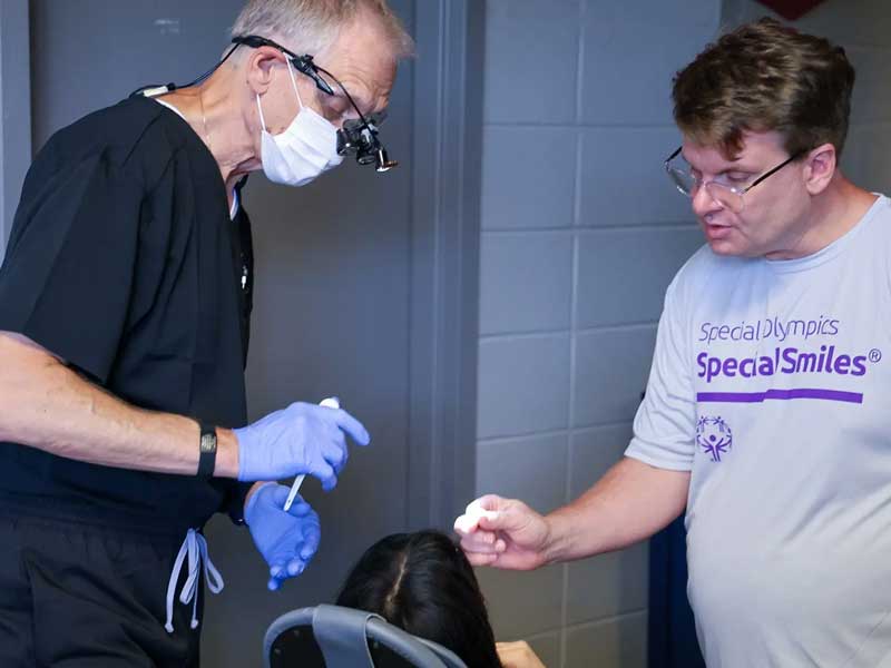 Two men standing over a person in a dental chair. One is wearing dental protective equipment. The other is wearing a t-shirt that says \