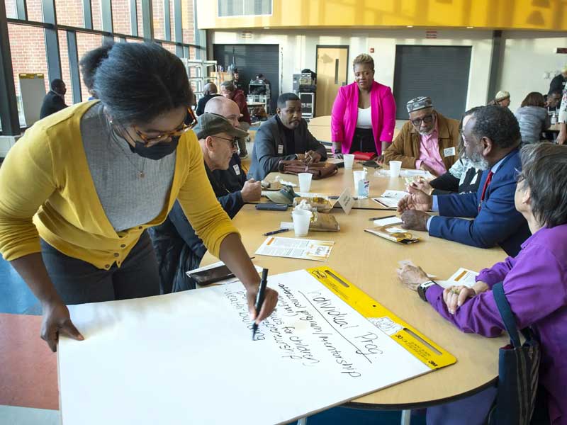 A person at a meeting is writing on a giant pad of paper while a group of community members is gathered around a table