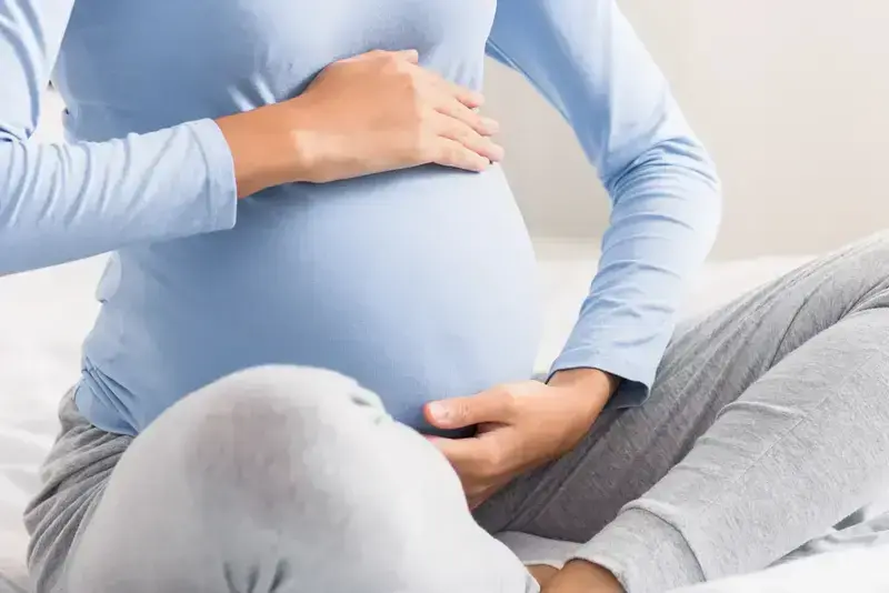 A photo of a woman from the neck down. She is sitting with her legs crossed and her hands cupping the top and bottom of her pregnant stomach.