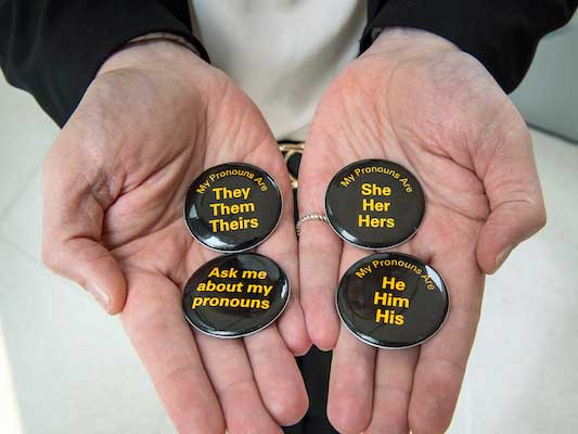 a pair of hands displaying four lapel pins that can be worn to denote what pronouns the wearer prefers